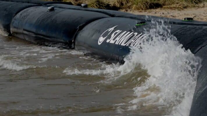 A black water-filled flood barrier labeled "SAMMYS" blocks and diverts waves along a shoreline as water splashes against it.