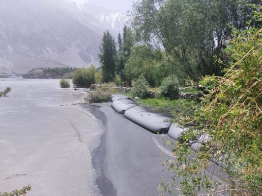A riverbank with black sandbags lining the shore, surrounded by green trees and mountains in the background—an inspiring scene from a Climate Innovation Challenge winner’s project.