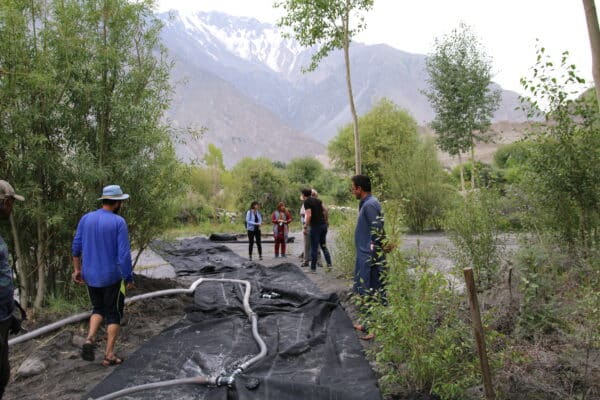Several people, including a Climate Innovation Challenge winner, stand and walk on black tarps and hoses laid on the ground in a green, mountainous outdoor area, with trees and mountains in the background.