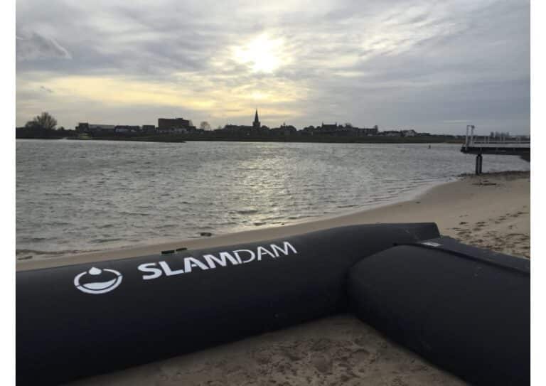 A SLAMDAM flood barrier, part of integrated water management, is positioned on a sandy beach beside a river, with a town and cloudy sky visible in the background at sunset.