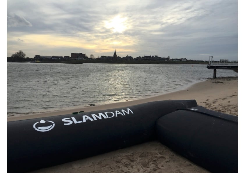 A SLAMDAM flood barrier, part of integrated water management, is positioned on a sandy beach beside a river, with a town and cloudy sky visible in the background at sunset.