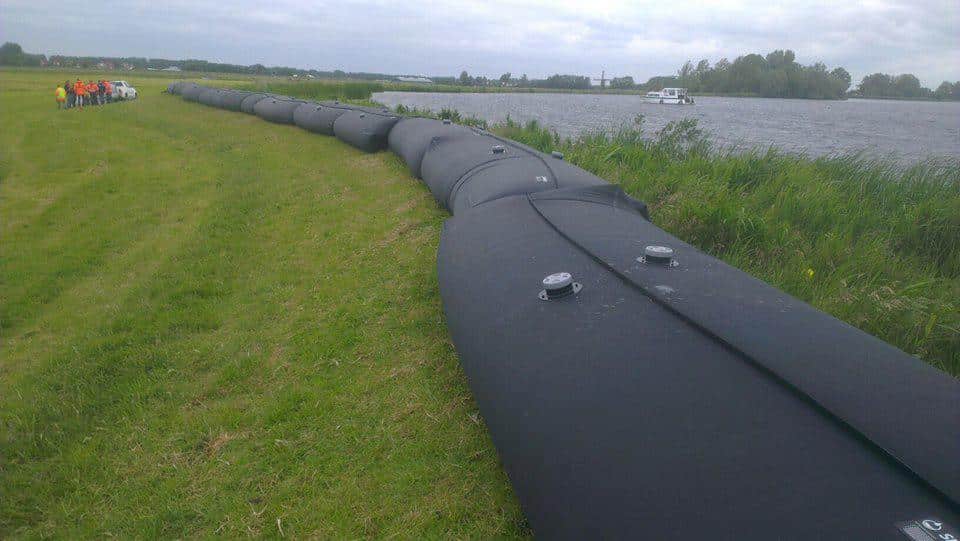 Large black water-filled flood barrier runs along a grassy riverbank, showcasing integrated water management, with a group of people and a vehicle visible in the distance.