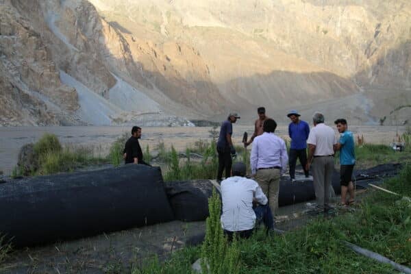 A group of people, including a Climate Innovation Challenge winner, stand and talk near a large black pipe or tube in a grassy area with rocky mountains and a river in the background.
