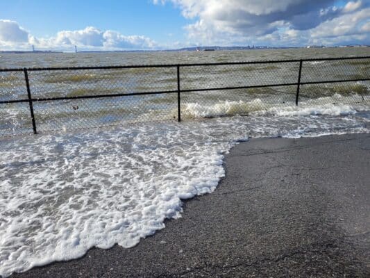 Ocean waves washing over a paved walkway, with a metal fence and cloudy sky in the background, capturing the environmental challenges highlighted by the Governors Island Climate Solutions Challenge.