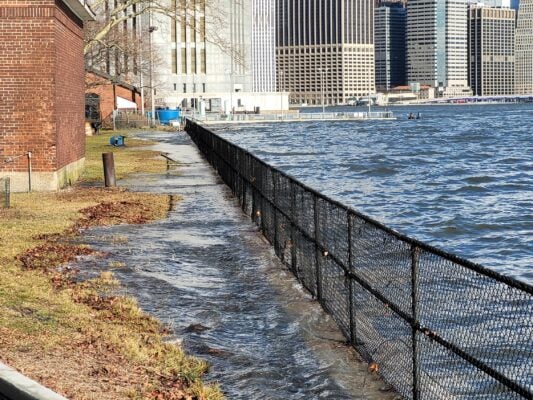 Floodwater rises onto a grassy path next to a black chain-link fence along a city waterfront, with tall buildings in the background—a striking scene reminiscent of challenges addressed by the Governors Island Climate Solutions Challenge.