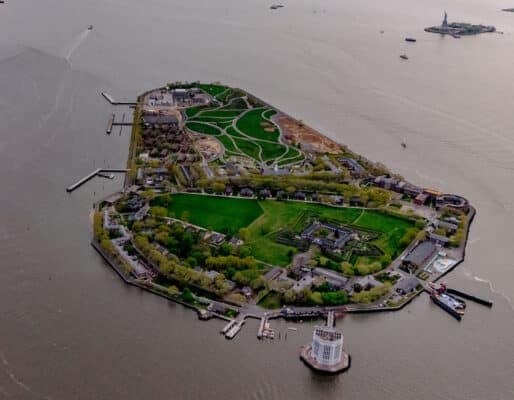 Aerial view of Governors Island in New York Harbor, showing green lawns, historic buildings, and surrounding water. The site is a focal point for the Governors Island Climate Solutions Challenge, with the Statue of Liberty visible in the distance.