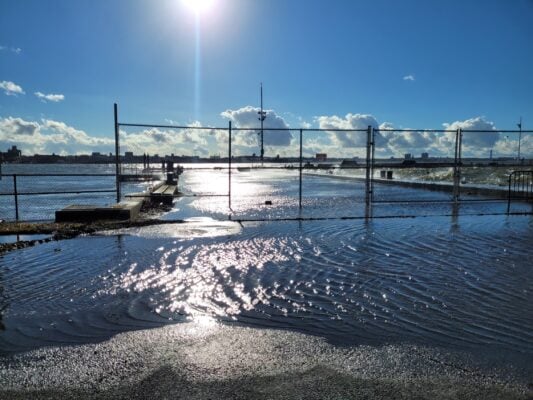 Flooded waterfront area with sunlight reflecting off water, a chain-link fence in the foreground, and city buildings visible in the distance under a clear blue sky—capturing the kind of resilience and adaptation envisioned by the Governors Island Climate Solutions Challenge.