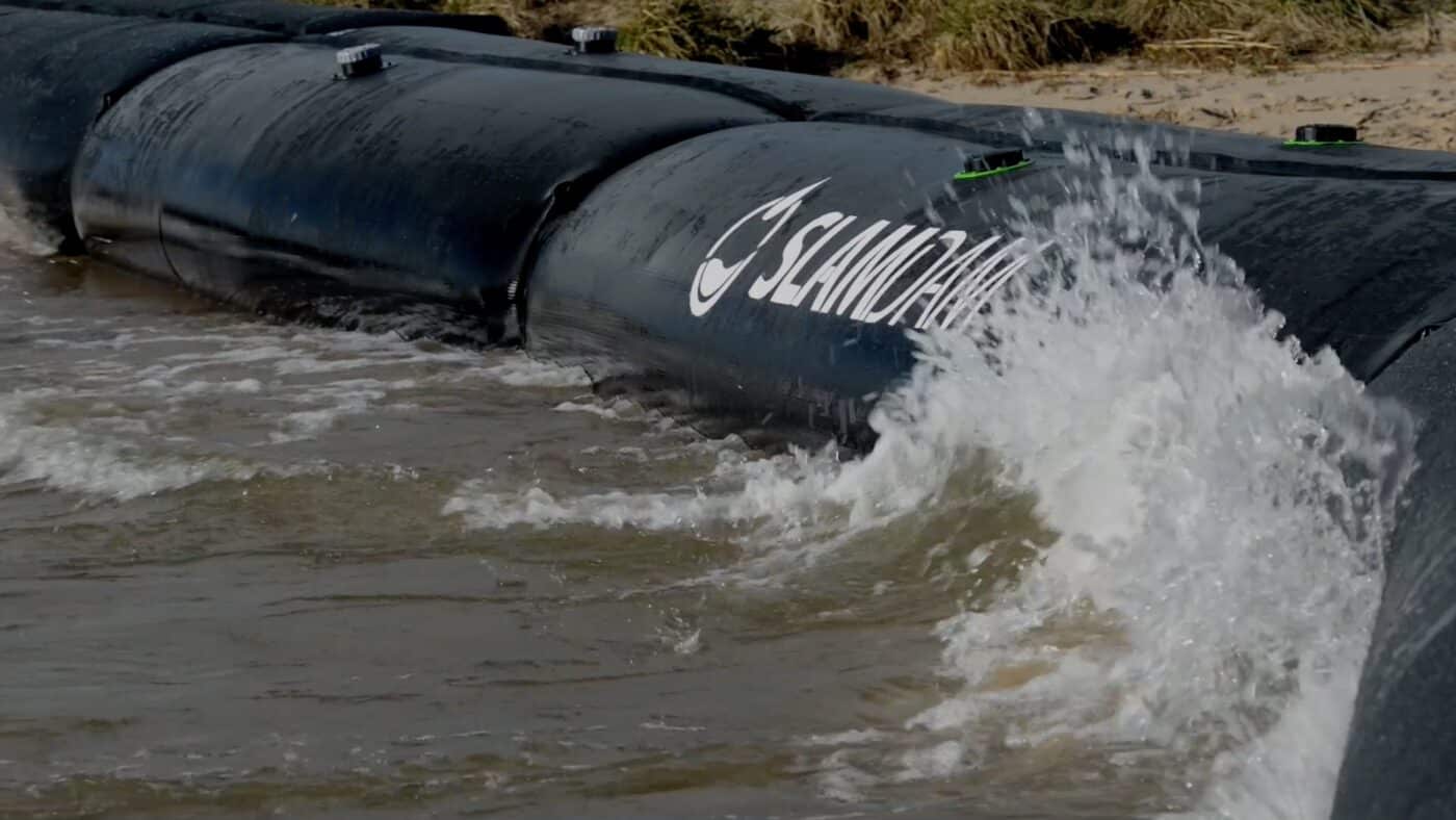 Large black water-filled barrier labeled "Sandbaggy" blocks moving water near a shoreline on Governors Island, with waves splashing against it—a scene reminiscent of innovations showcased in the Governors Island Climate Solutions Challenge.
