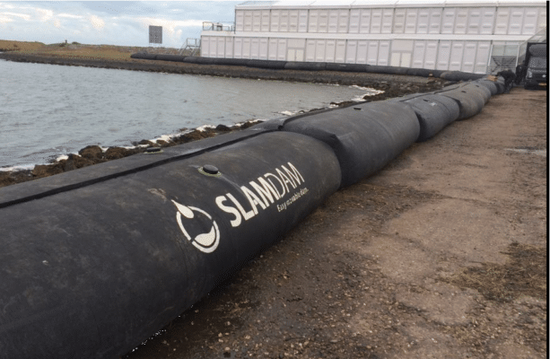 Large black water-filled flood barriers labeled "SLAMDAM" are placed along a shoreline near a white temporary structure, serving as an innovative flood defense demonstration for Climate Week NYC 2026.