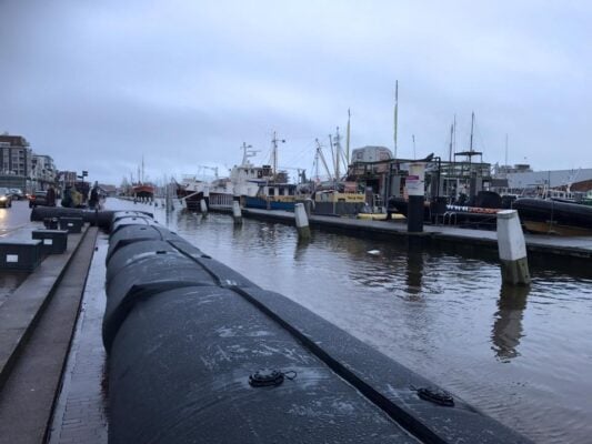 Several boats are docked in a harbor on a cloudy day, with large black flood barriers lining the waterfront street—a striking reminder of the city's resilience and adaptation efforts highlighted during Climate Week NYC 2026.